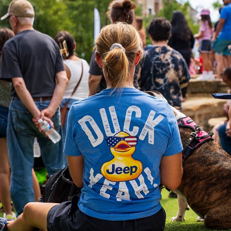 A woman with a ponytail wears a blue T-shirt with a cartoon duck, an American flag, and the words DUCK YEAH! and Jeep as she sits outdoors among a crowd; a dog in a pink harness is beside her.