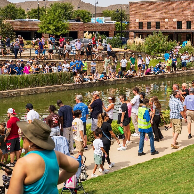 A large crowd of people gathers along both sides of a canal in a park on a sunny day, watching and taking photos. Some people are standing, others are sitting on the grass or stone wall near the water.