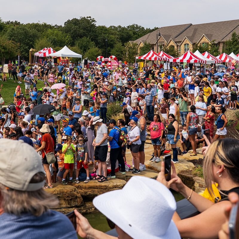 A large crowd gathers outdoors at a festival with red-and-white striped tents, food stalls, and people taking photos near a pond on a sunny day. Some wear hats, and children and families are present.