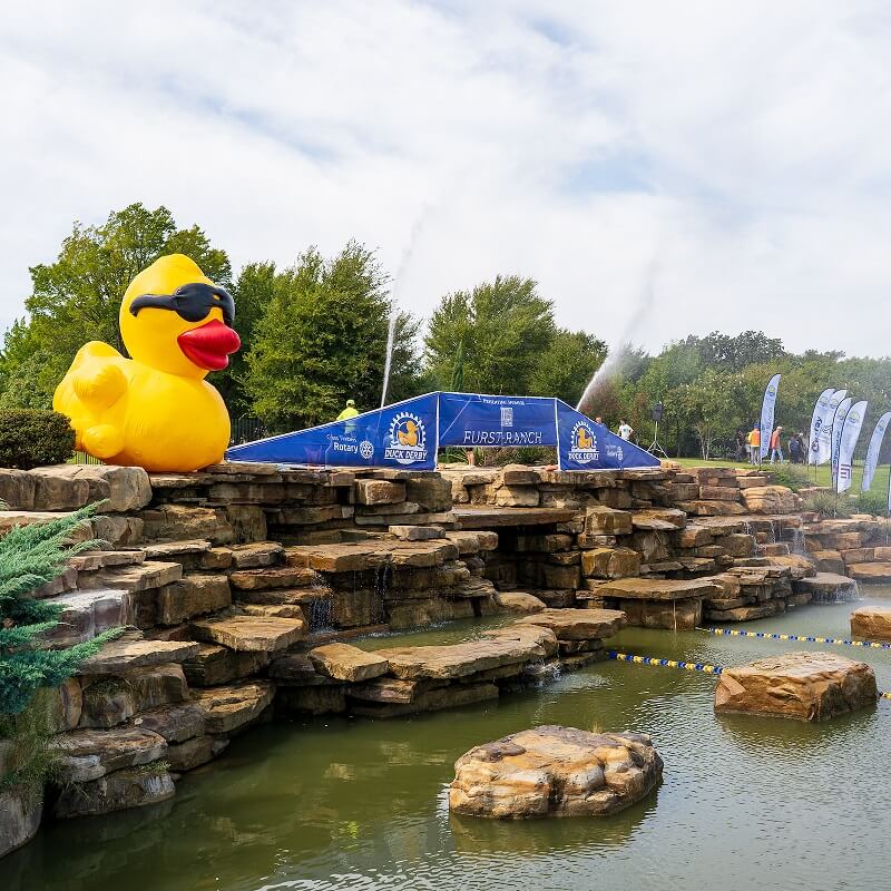 A large inflatable yellow duck wearing sunglasses sits on rocks by a pond with water fountains and greenery in the background; event banners are displayed behind the duck.
