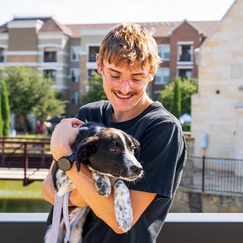 A young man with glasses and a mustache smiles while holding a black and white puppy outside, with buildings and trees in the background.